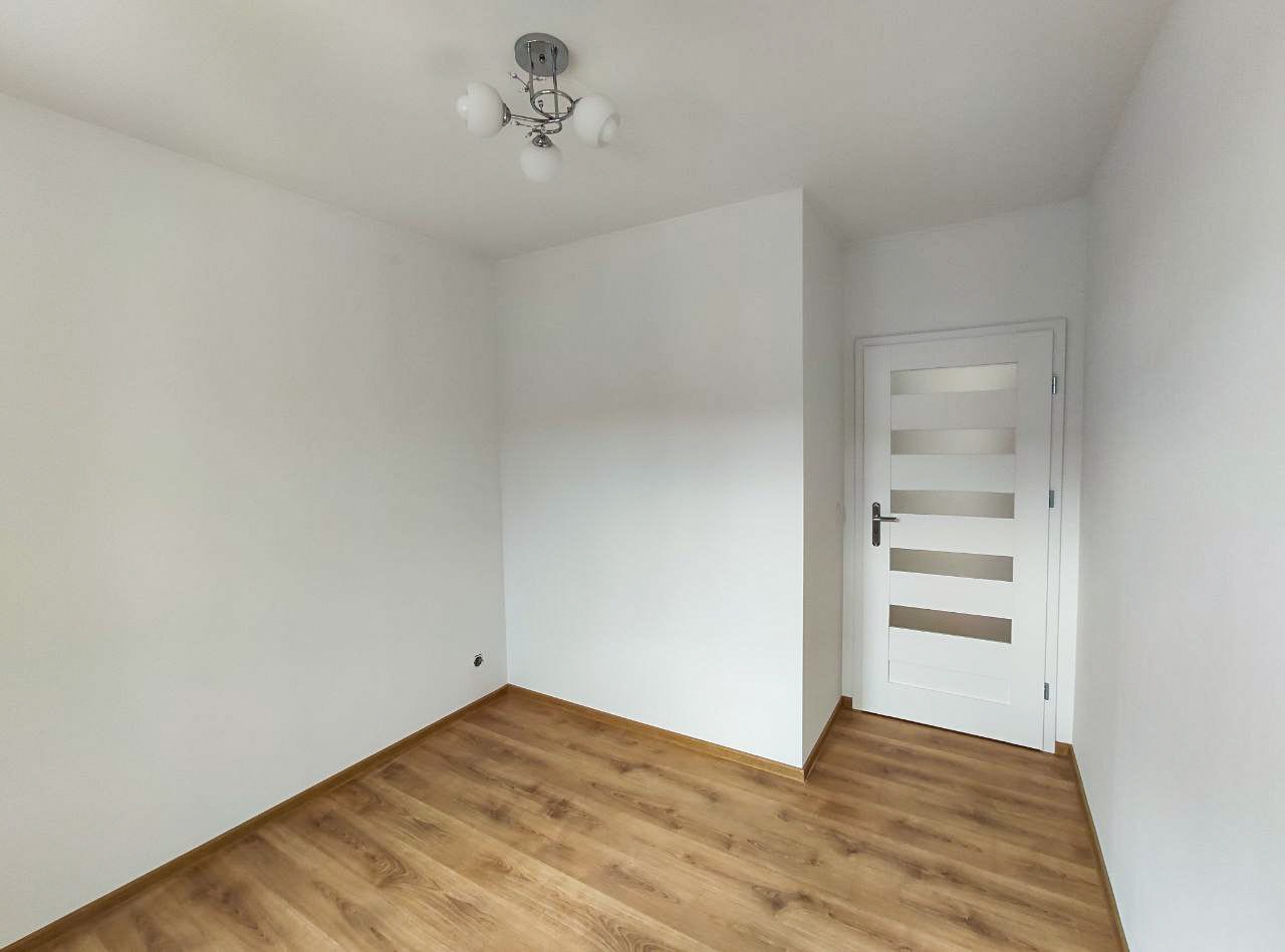 Pusty pokój z drewnianą podłogą z paneli, białymi ścianami i białymi drzwiami z poziomymi przeszkleniami, oświetlony przez nowoczesną lampę sufitową.