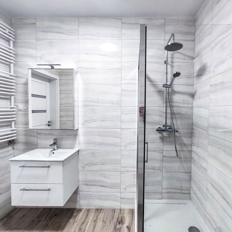 Nowoczesna łazienka z kabiną prysznicową, umywalką z szafką i grzejnikiem drabinkowym. Ściany wyłożone płytkami imitującymi marmur, podłoga z paneli drewnopodobnych.