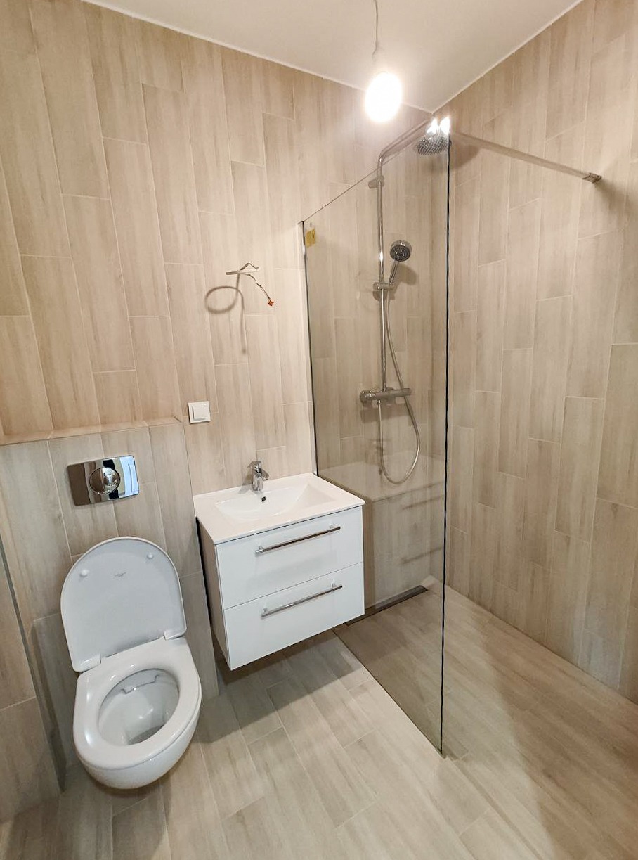 Nowoczesna łazienka z beżowymi płytkami, kabiną prysznicową typu walk-in, podwieszaną szafką z umywalką i toaletą z otwartą klapą.