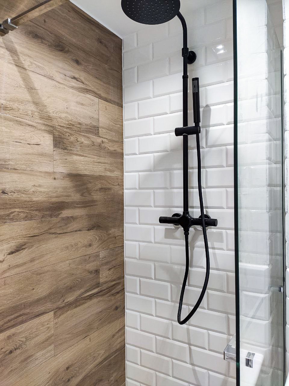 Nowoczesna kabina prysznicowa z czarną armaturą na tle białych, prostokątnych płytek typu metro i drewnopodobnych paneli ściennych.