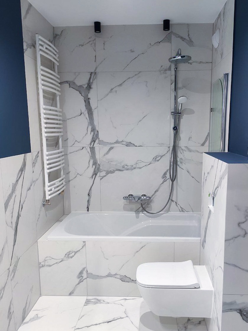 Nowoczesna łazienka z wanną obudowaną płytkami imitującymi marmur, wiszącą toaletą i grzejnikiem drabinkowym, oświetlona punktowymi lampami sufitowymi, z fragmentem ściany w kolorze granatowym.