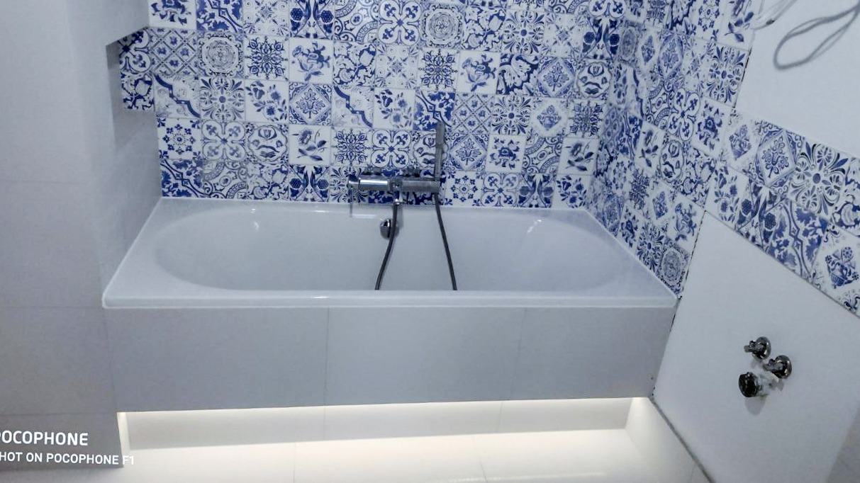 Biała wanna z chromowaną armaturą na tle ściany wyłożonej dekoracyjnymi, biało-niebieskimi płytkami. Podświetlenie LED pod wanną. Widoczne zawory na ścianie obok wanny.