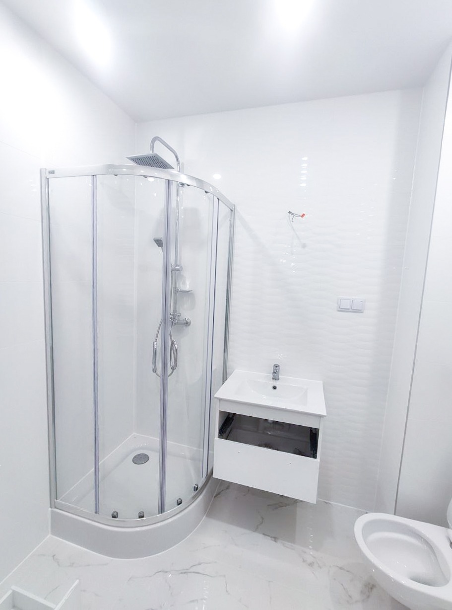 Jasna łazienka z kabiną prysznicową z chromowanymi elementami, białą umywalką z szafką bez frontów i toaletą, wykończona białymi płytkami imitującymi marmur.