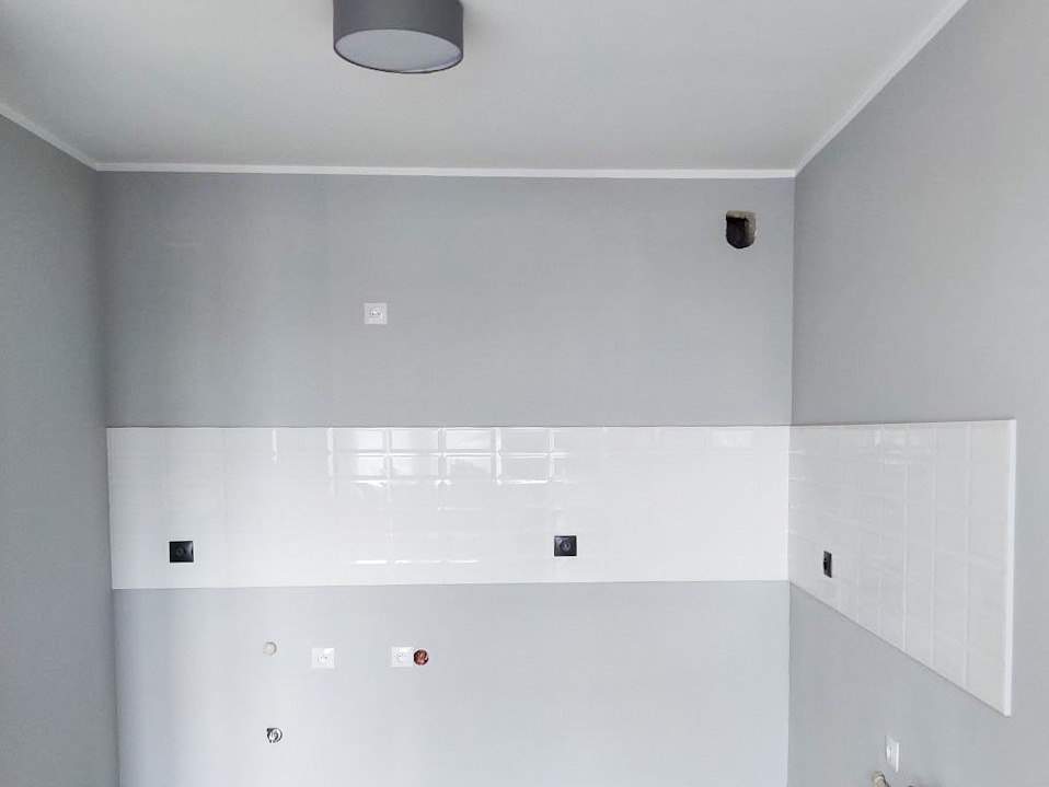 Świeżo ułożone, błyszczące, białe płytki ceramiczne na szarej ścianie w kuchni, widoczne przygotowane otwory na gniazdka elektryczne, surowy stan wykończenia.