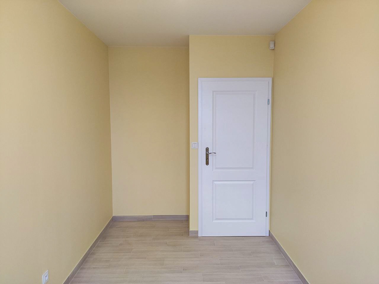 Pomalowane na jasnożółto puste pomieszczenie z białymi drzwiami i panelami podłogowymi imitującymi drewno, widoczny fragment sufitu i listwy przypodłogowe.