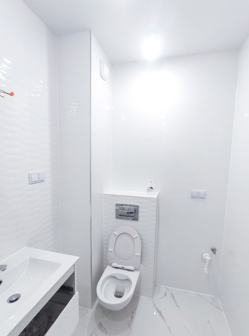 Nowoczesna, minimalistyczna łazienka z białymi, teksturowanymi płytkami na ścianach, podwieszaną toaletą z chromowanym przyciskiem spłukiwania, białą umywalką z szafką i marmurową podłogą...