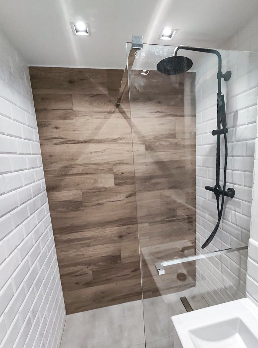 Nowoczesna kabina prysznicowa ze szklaną ścianą, czarną armaturą i drewnopodobną okładziną ścienną, oświetlona punktowymi lampami sufitowymi.