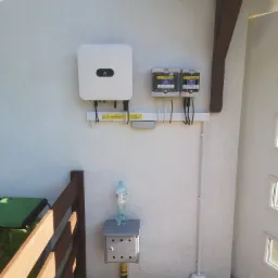 Zamontowany inwerter fotowoltaiczny Huawei i rozdzielnice PV na elewacji budynku, widoczne przewody, rurki instalacyjne, skrzynka gazowa z butelką wody na górze i fragment drewnianego ogrodzenia.