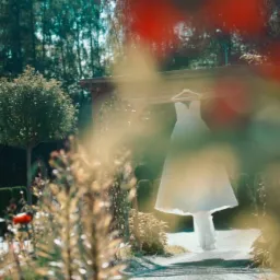 Suknia ślubna zwieszona na wieszaku z drewnianej altany, widoczna przez rozmyte kwiaty i zieleń ogrodu w słoneczny dzień.