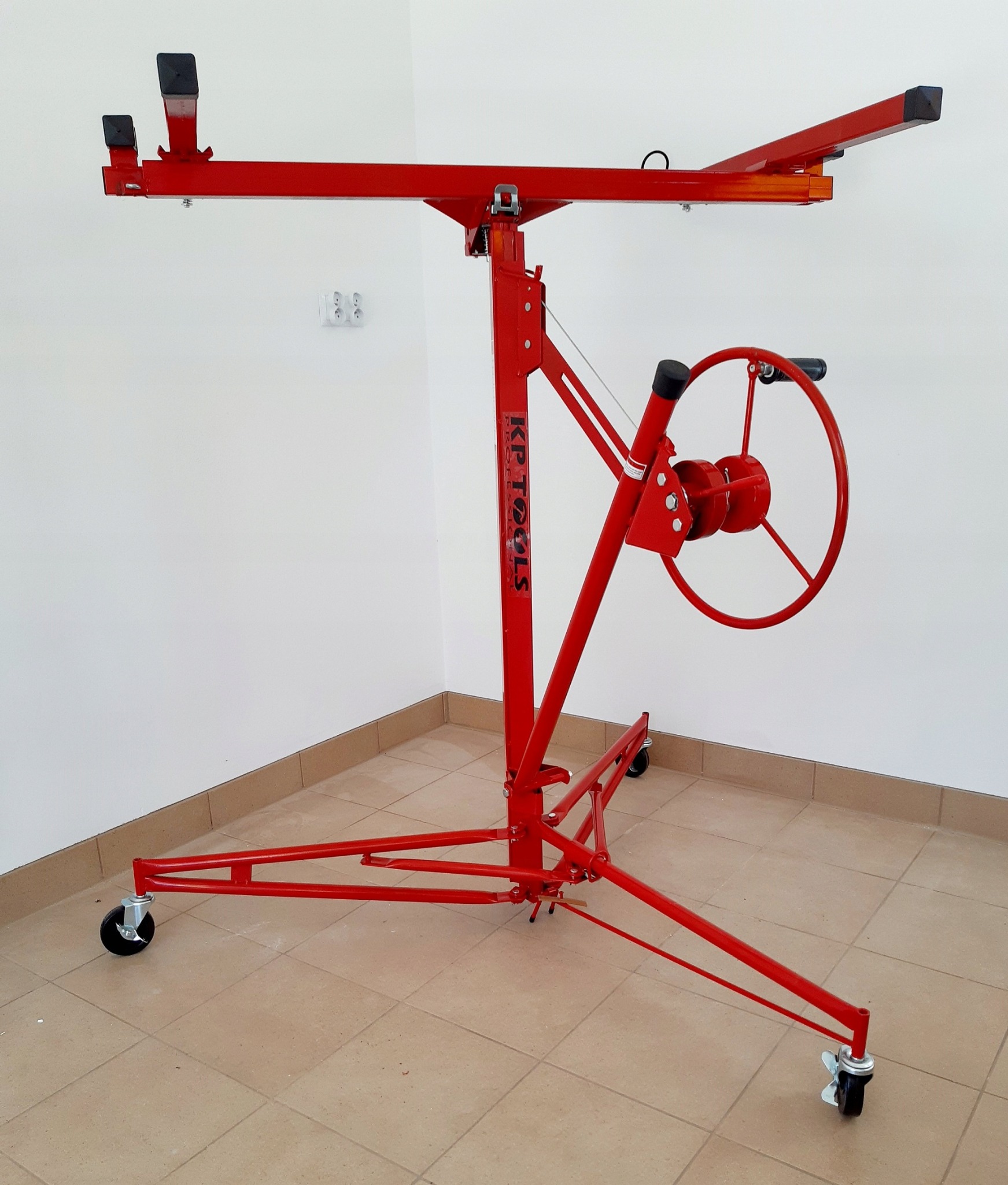 Czerwony podnośnik do płyt gipsowo-kartonowych marki KP TOOLS, widoczny w całości na tle białej ściany i beżowej podłogi z płytek, z kołami u podstawy i korbą do podnoszenia.