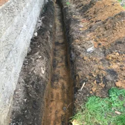 Wąski wykop wzdłuż fundamentu budynku z bloczków betonowych, ziemia usypana po bokach, koparka widoczna w tle.