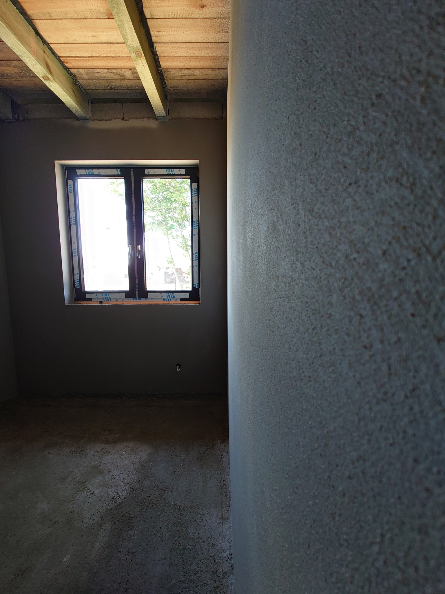 Wnętrze surowego pokoju z oknem, ściany pokryte świeżym, szarym tynkiem, widoczne drewniane belki stropowe, podłoga betonowa w trakcie prac wykończeniowych.