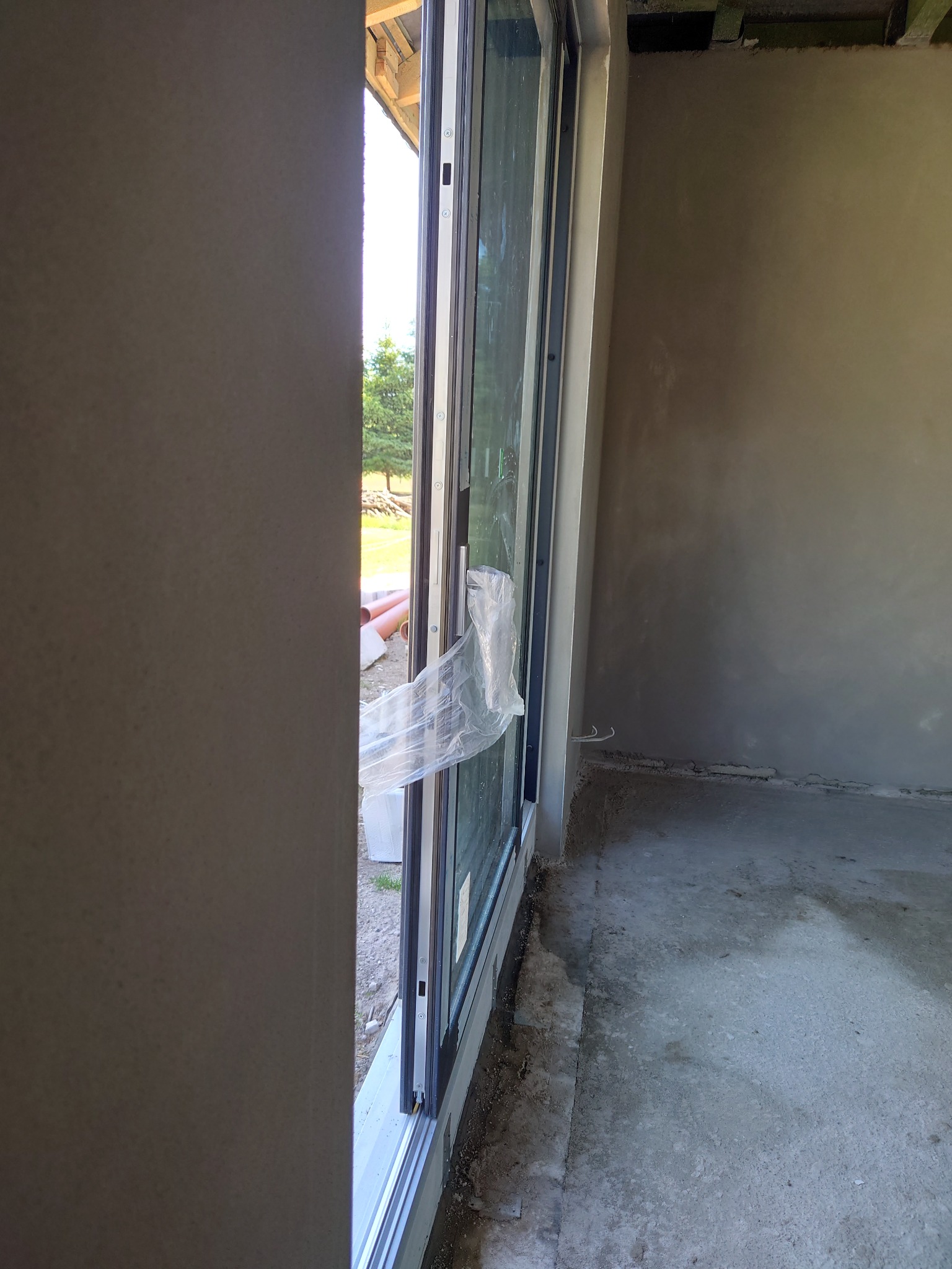 Świeżo nałożony, gładki tynk na ścianie wewnętrznej w surowym stanie, widok przez częściowo osłonięte folią okno przesuwne na zewnątrz.