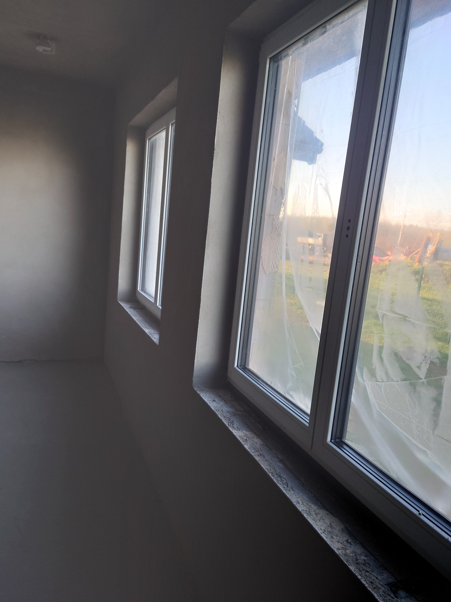 Wnętrze po nałożeniu gładzi szpachlowej, widoczne dwa białe okna zabezpieczone folią, jasne ściany w odcieniach szarości.