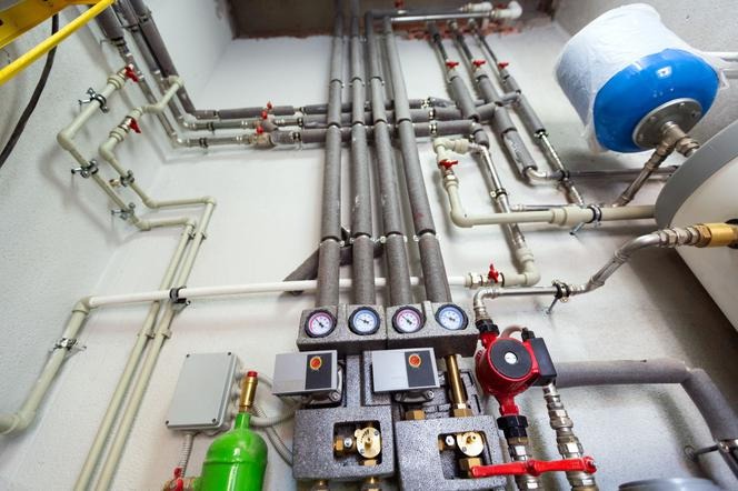 Złożona instalacja grzewcza z rurami o różnej średnicy, zaworami z czerwonymi pokrętłami, manometrami i pompą obiegową, całość umieszczona na białej ścianie.