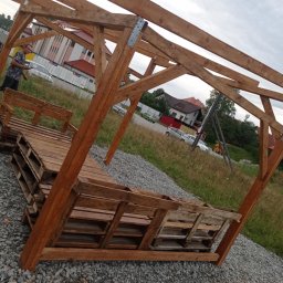 F.H.U. Kacper Kapłon - Konstrukcja altany z palet na żwirowym podłożu, widoczny fragment budynku w tle i osoba po lewej stronie. Drewno impregnowane, surowy styl.