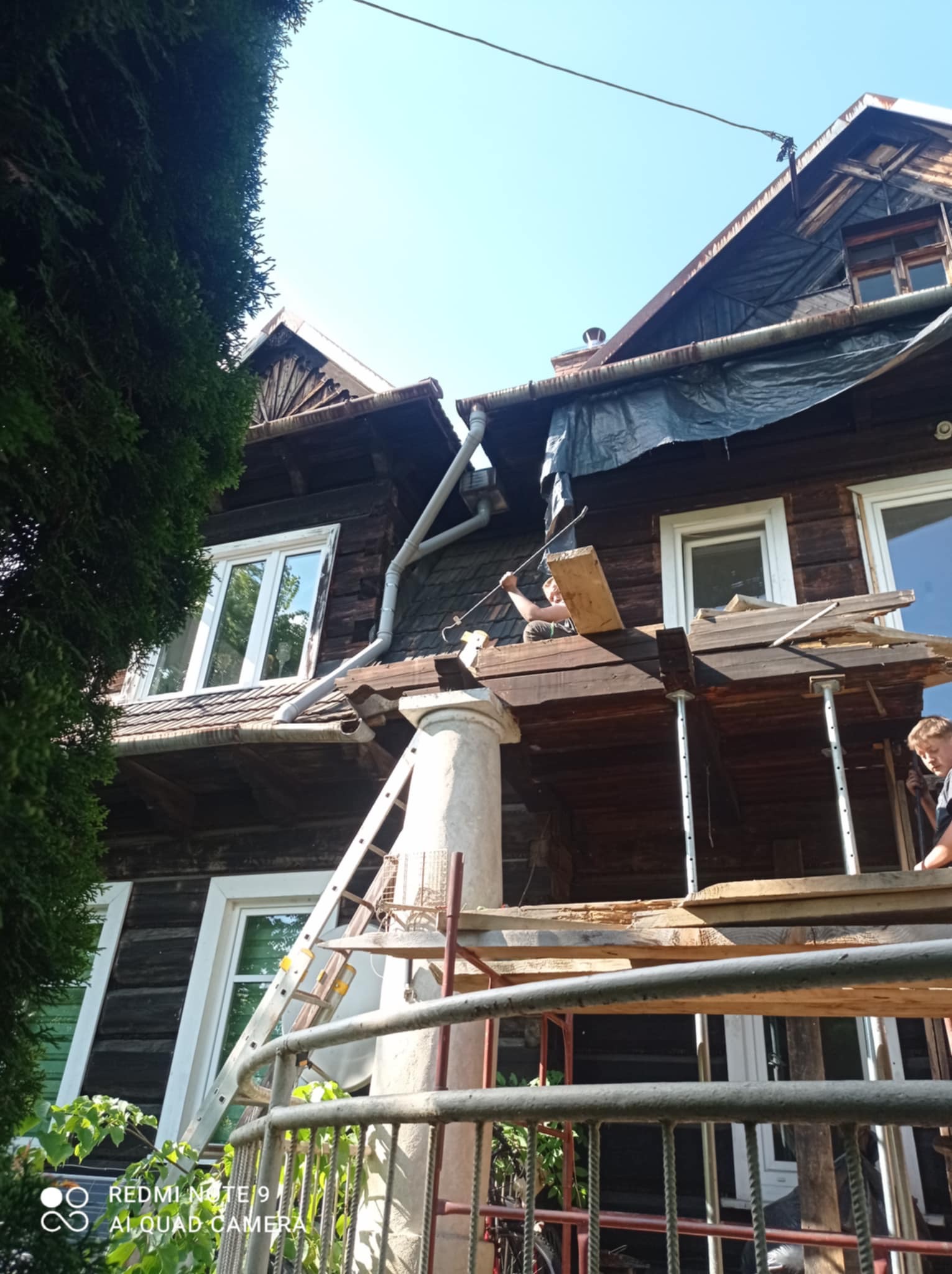 Renowacja drewnianego domu z rusztowaniem i drabina. Widoczne elementy konstrukcyjne, okna i pracownicy podczas prac remontowych. Częściowo zakryty dach folią.