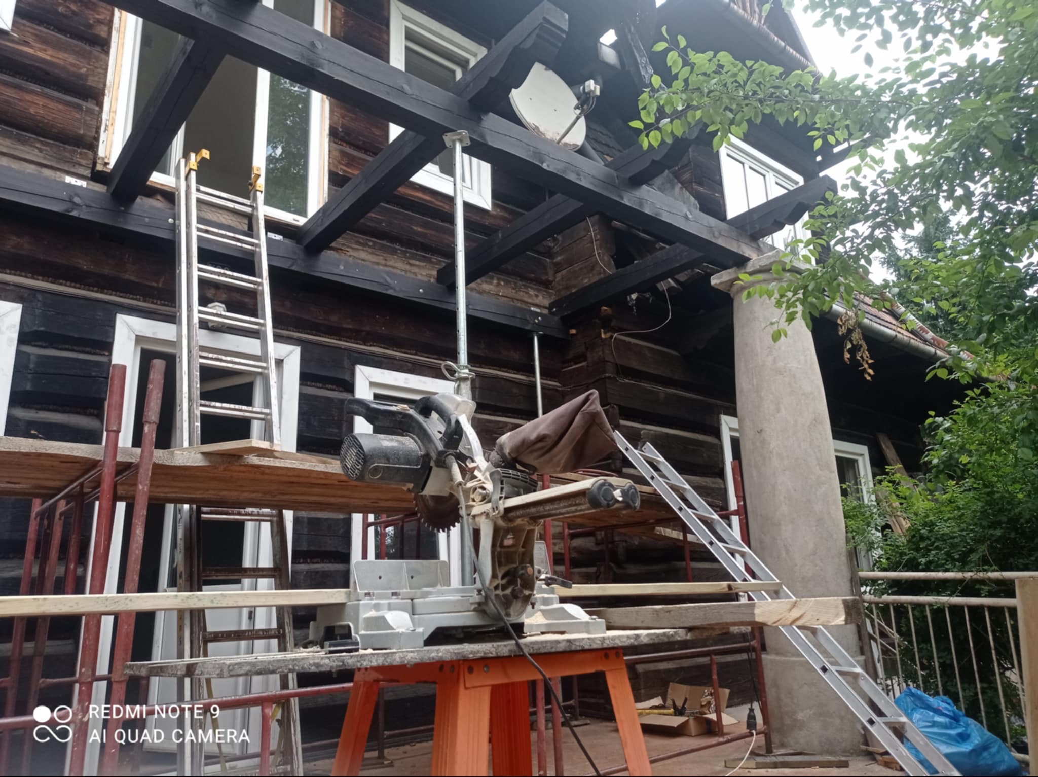 Budowa drewnianego domu z bali: piła stołowa na pierwszym planie, rusztowania i drabiny przy ścianie z oknami. Widoczna konstrukcja dachu i satelita.