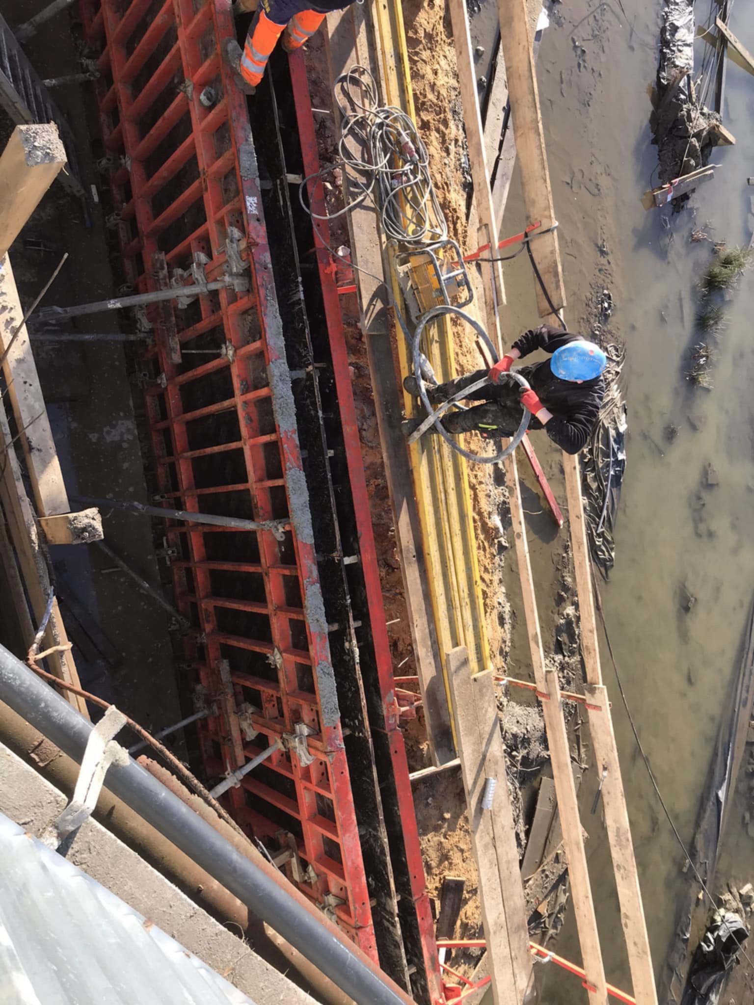 Z góry widok na pracownika w kasku, który pracuje przy szalunku fundamentów, z widocznym zbrojeniem i częściowo zalanym wykopem. Prace ziemne w toku.