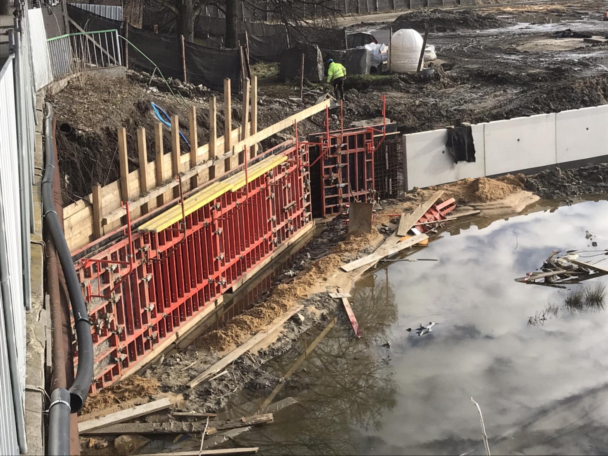 Budowa żelbetowej konstrukcji oporowej w wykopie z częściowym zalaniem wodą. Widoczne szalunki, zbrojenia i pracownik w odblaskowej kurtce.