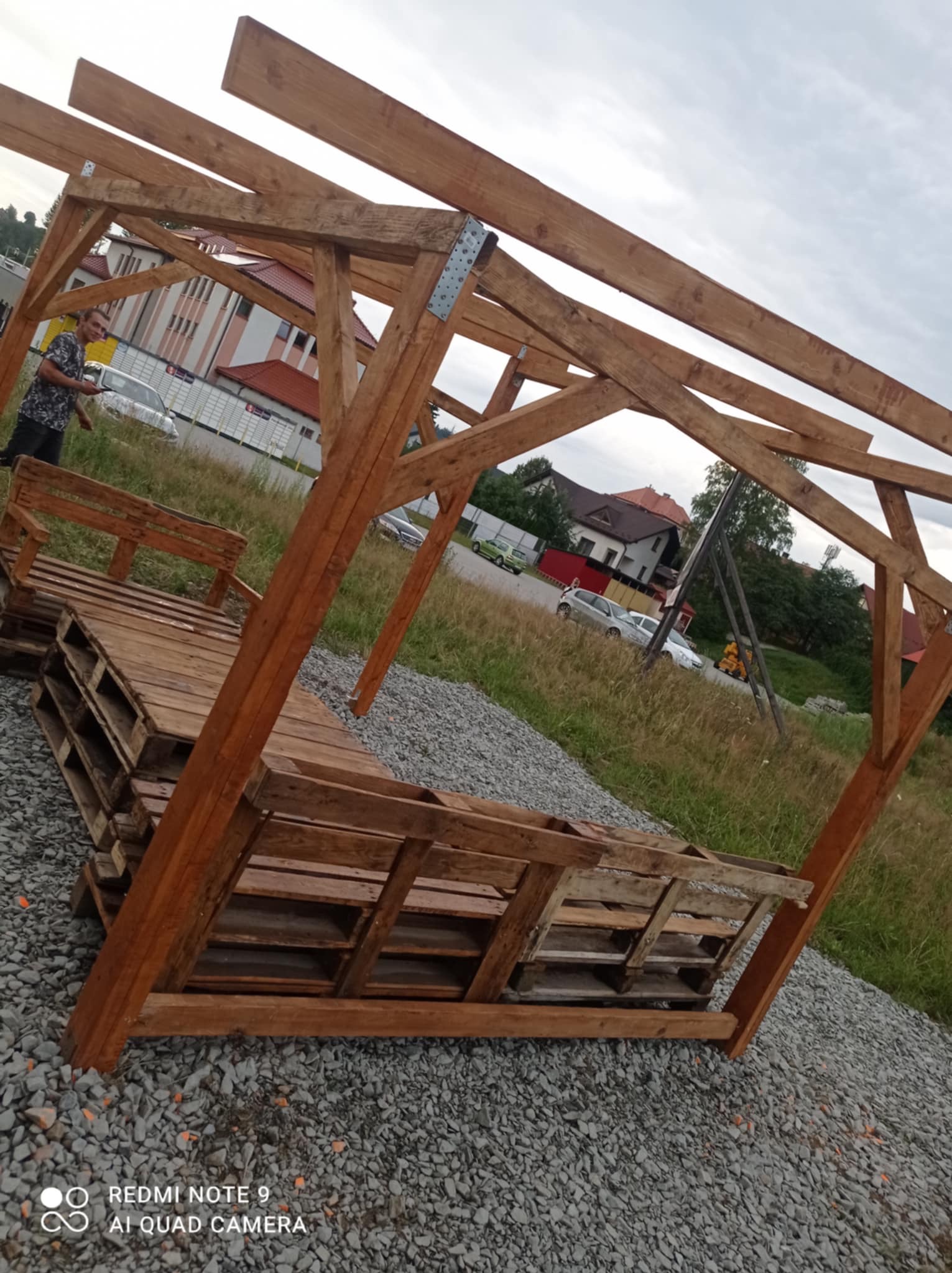 Konstrukcja altany z palet na żwirowym podłożu, widoczny fragment budynku w tle i osoba po lewej stronie. Drewno impregnowane, surowy styl.