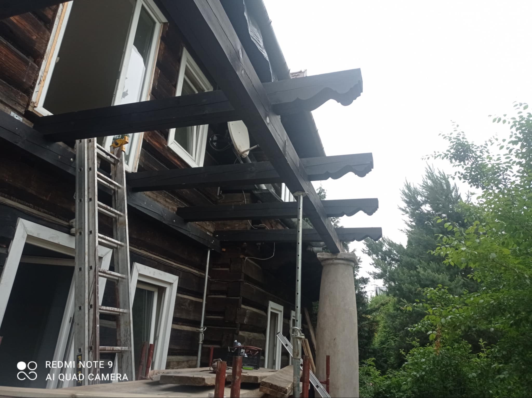 Renowacja drewnianego domu z nową konstrukcją zadaszenia, widoczne rusztowania i drabina. Detal: dekoracyjne zakończenia belek zadaszenia.