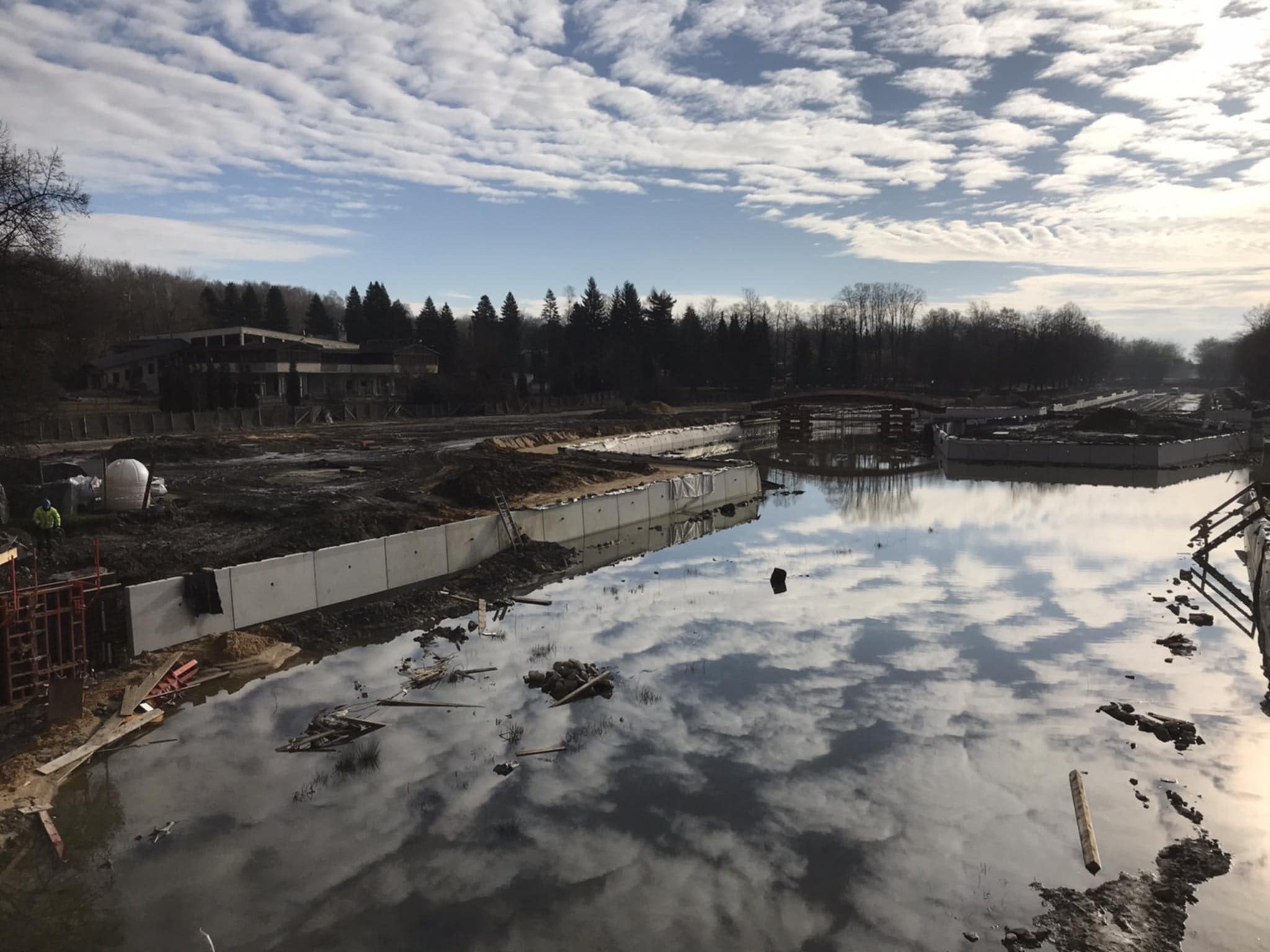Budowa zbiornika wodnego z betonowymi ścianami, odbicie chmur w wodzie, widoczny teren budowy i las w tle. Prace ziemne w toku, konstrukcja w trakcie realizacji.