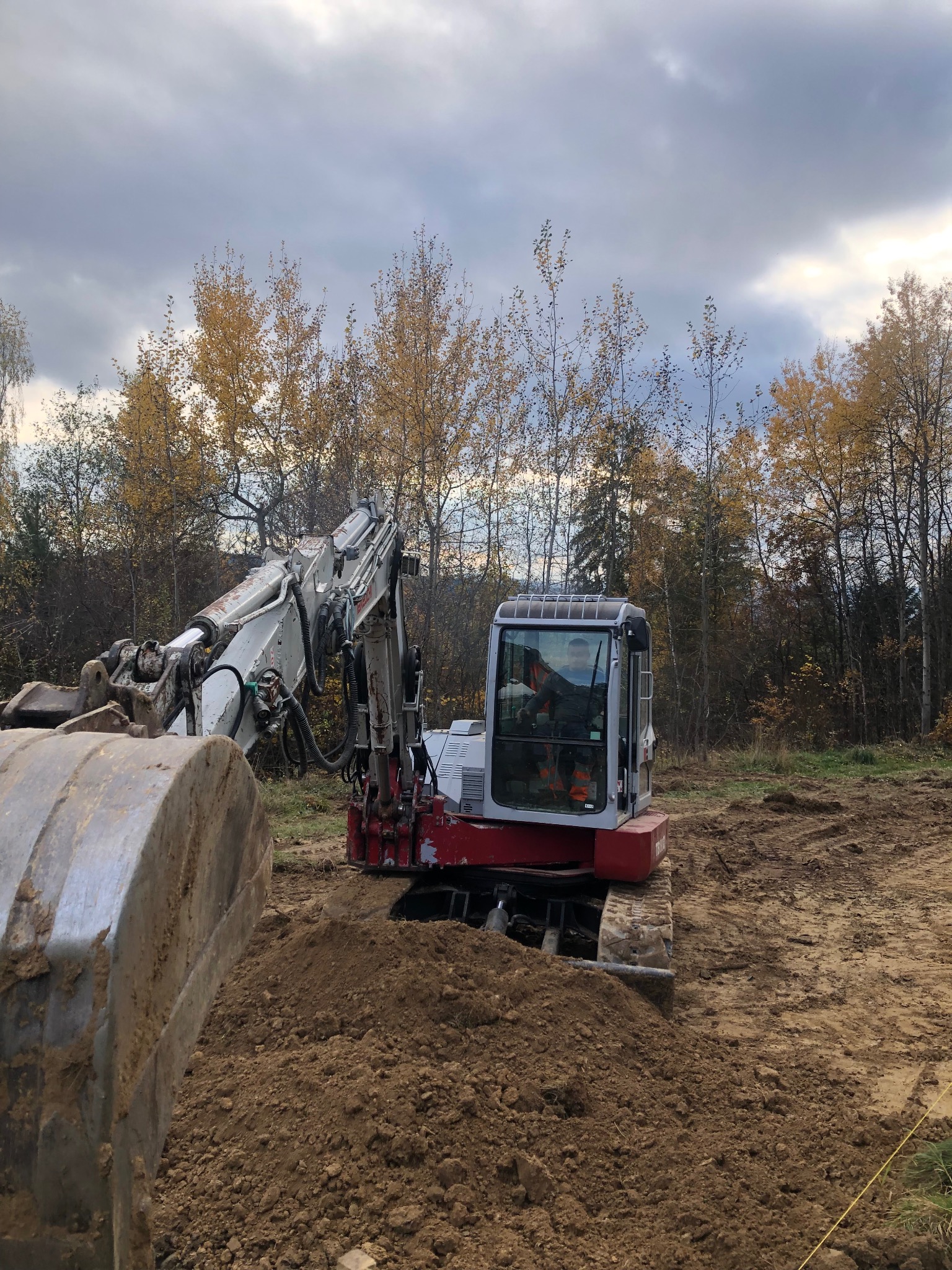 Biało-czerwona koparka gąsienicowa w trakcie prac ziemnych na działce z drzewami w tle, widok z bliska na łyżkę koparki.