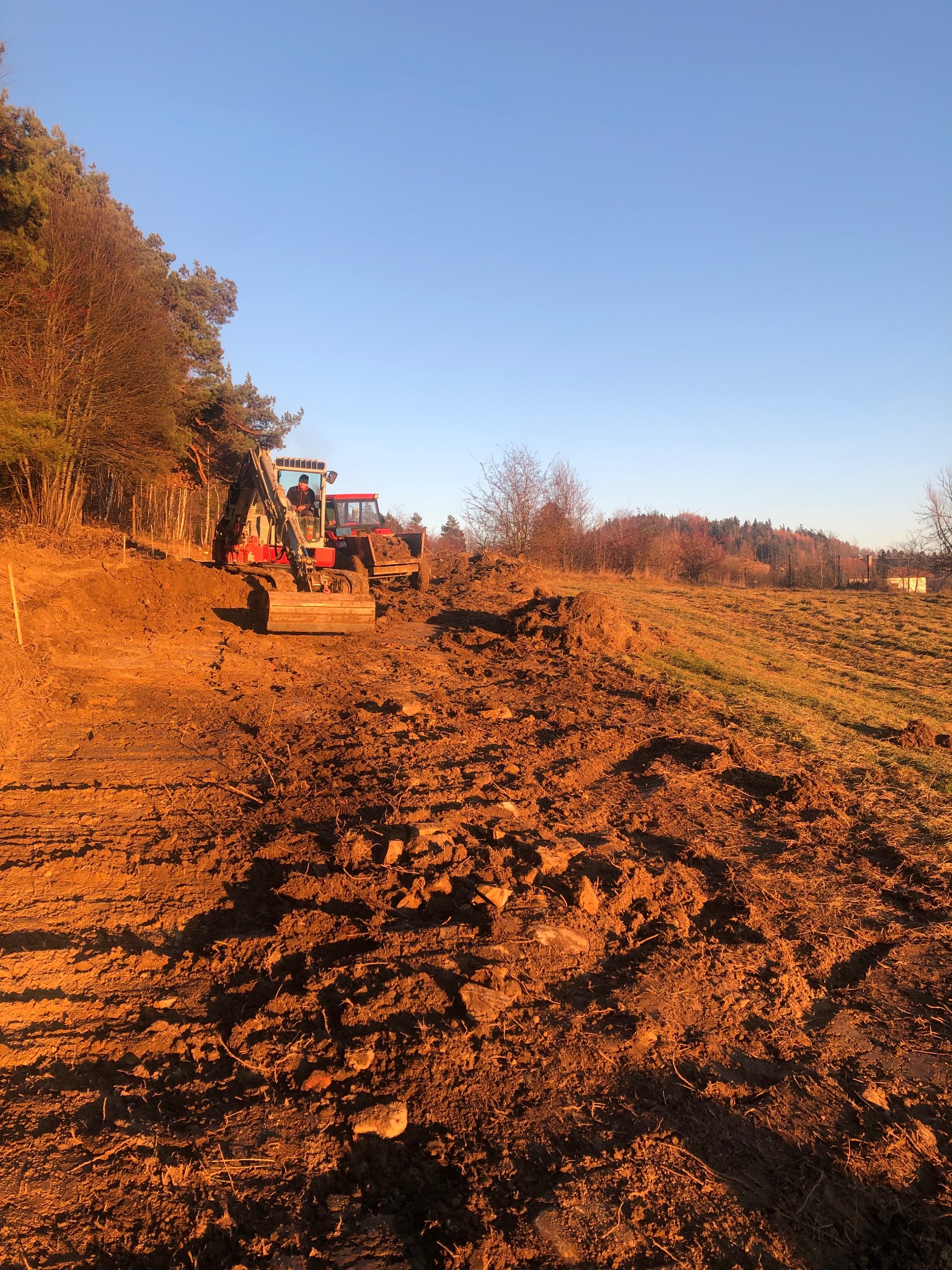 Czerwony traktor i koparka na nierównym, brązowym terenie w słoneczny dzień. W tle las i błękitne niebo. Prace ziemne w toku.