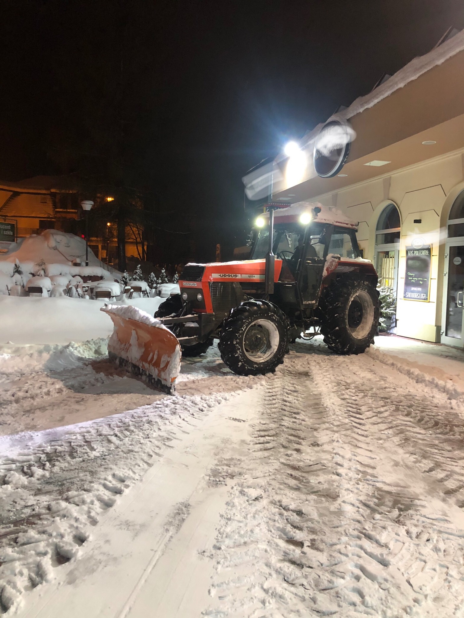 Czerwony traktor Ursus z pługiem odśnieża teren przed budynkiem w nocy. Widoczne ślady opon w śniegu i oświetlony budynek w tle.