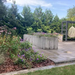 Nowoczesny ogród z betonowymi donicami wypełnionymi fioletowymi kwiatami, wyłożony płytami taras, z wiszącymi fotelami w kształcie łezki i żywopłotem z tui w tle.