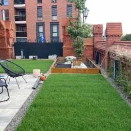 Nowo założony trawnik z rolki na tle ceglanego budynku z elementami dekoracyjnymi, widoczne narzędzia ogrodnicze, krzesła ogrodowe i przygotowane miejsce na nasadzenia.