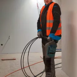 Uśmiechnięty elektryk w kasku z latarką i kamizelce odblaskowej trzyma wiązkę czarnych kabli na tle surowego wnętrza z widocznymi przewodami i ostrzeżeniem o gazie.