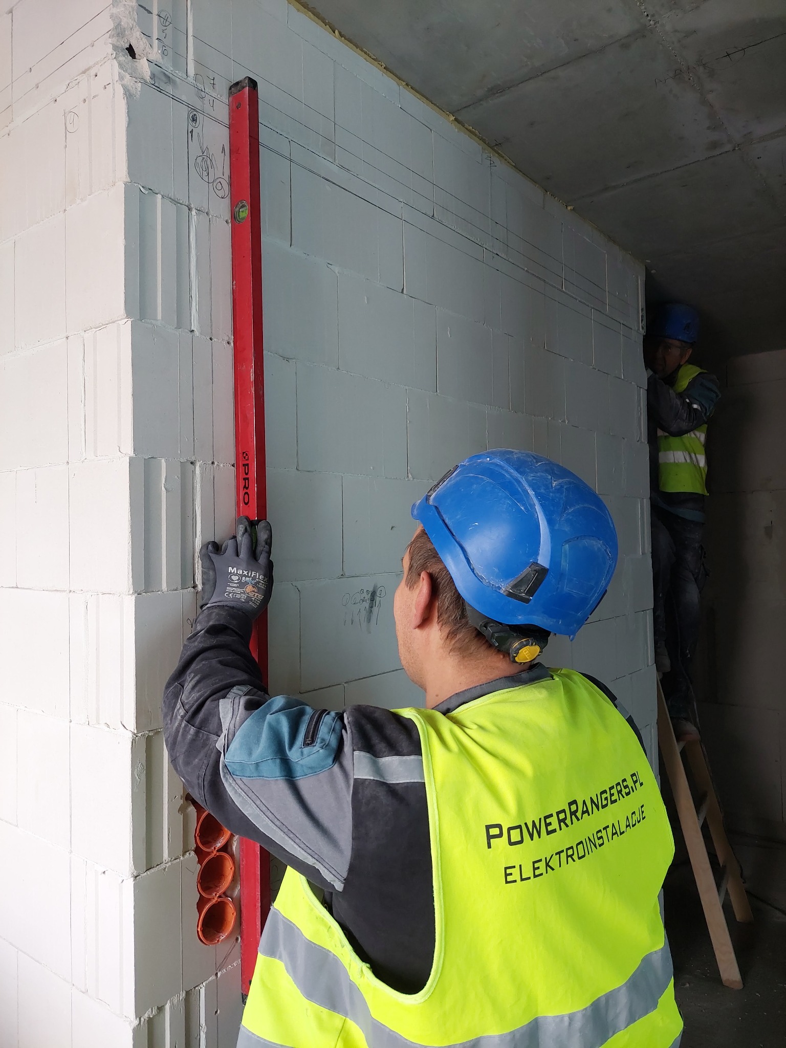 Elektryk w kamizelce odblaskowej z logo firmy PowerRangers.pl Elektroinstalacje, mierzy pion pionową poziomicą na ścianie z betonu komórkowego, na której zaznaczone są linie prowadzące pod kable;...