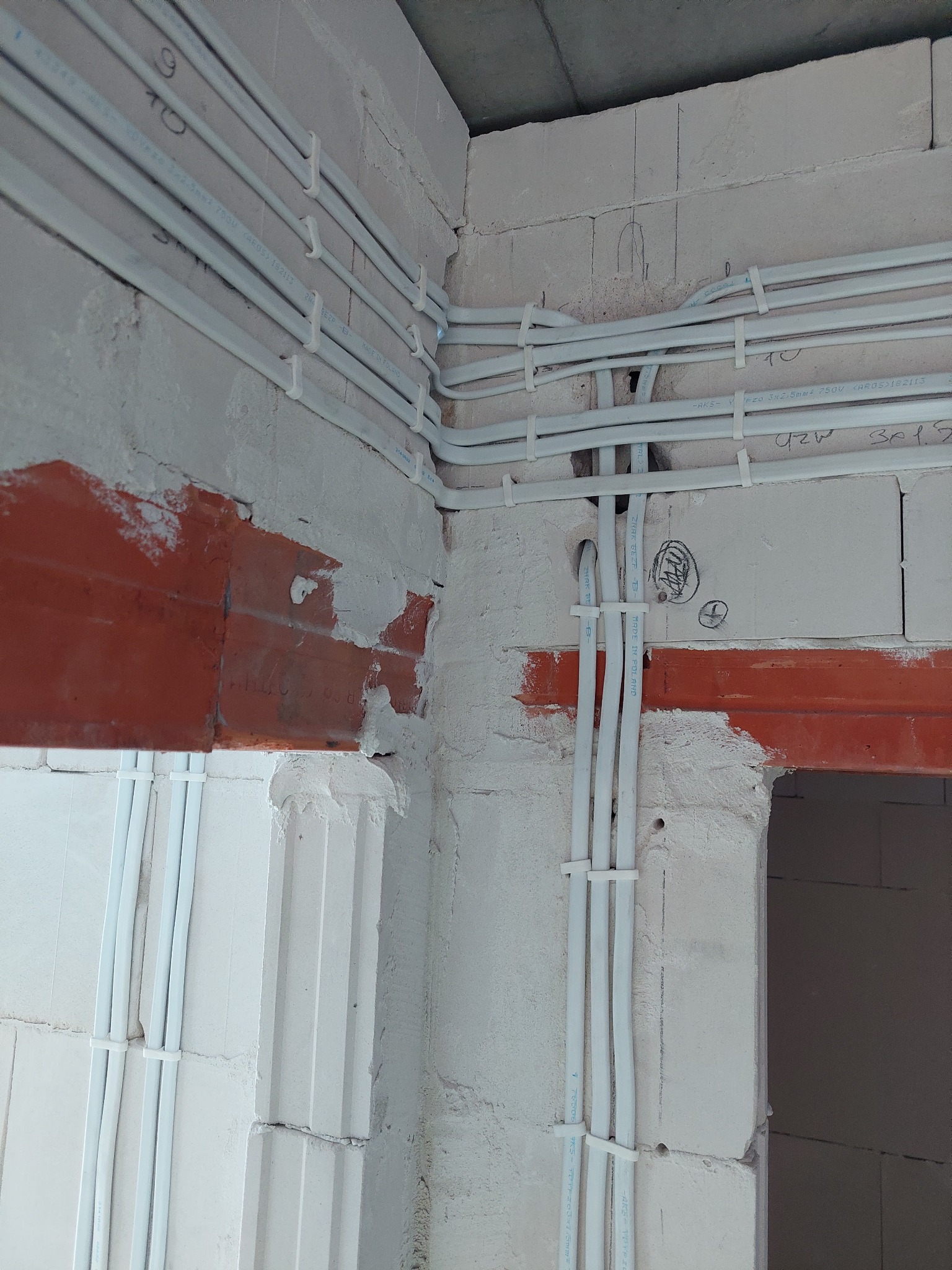 Białe kable elektryczne ułożone wzdłuż surowej ściany z betonu komórkowego, przymocowane plastikowymi uchwytami. Widoczne oznaczenia na kablach i ścianie, w tle otwór w ścianie.