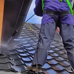 Pracownik w kombinezonie i uprzęży, myjący czarny dach z dachówki ceramicznej myjką ciśnieniową, widok od dołu z perspektywy krzewów.