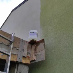 Fragment elewacji z widoczną różnicą w kolorze po częściowym czyszczeniu, na rusztowaniu stoi plastikowy pojemnik z preparatem czyszczącym.
