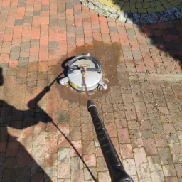 Czyszczenie kostki brukowej myjką ciśnieniową z okrągłą dyszą, widoczny cień osoby obsługującej urządzenie na mokrej powierzchni kostki.