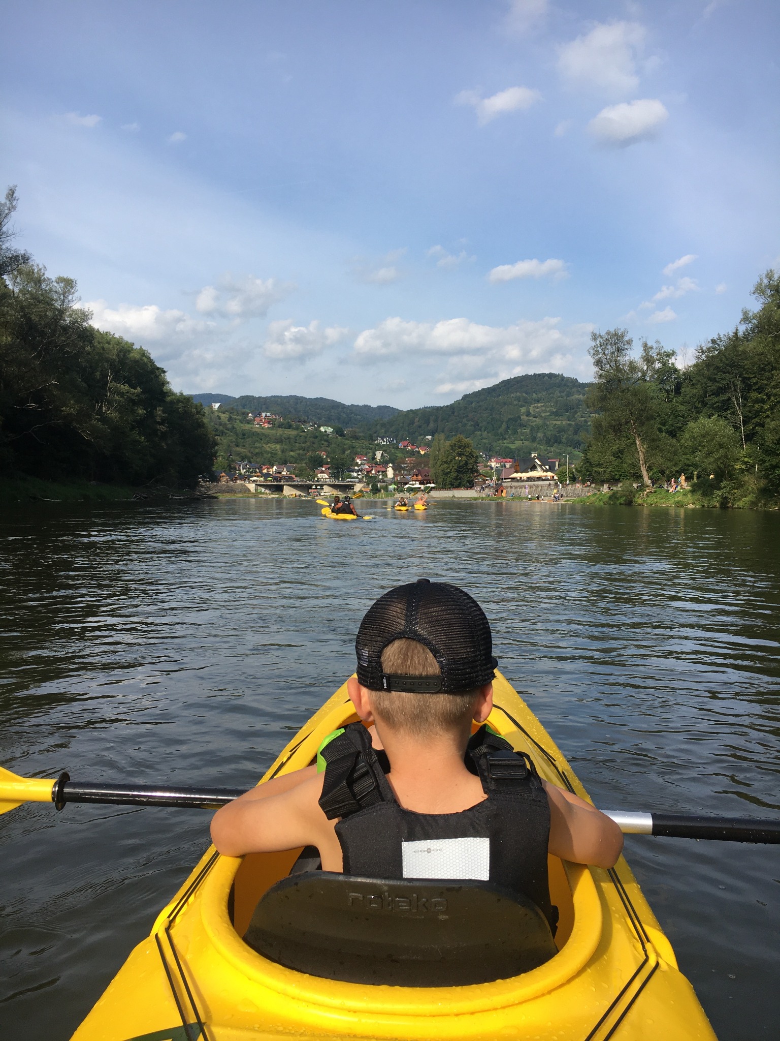 Chłopiec w czapce i kamizelce asekuracyjnej płynie kajakiem po rzece w kierunku miasteczka z widocznym mostem i innymi kajakami.