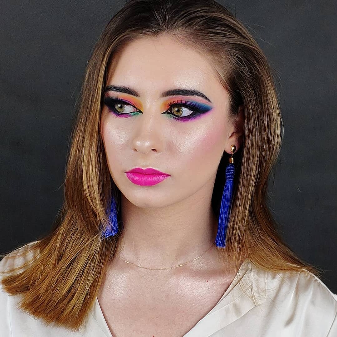 Portret kobiety z intensywnym, tęczowym cieniowaniem powiek i różową szminką, uzupełnionym długimi, niebieskimi kolczykami z frędzlami na tle ciemnoszarego tła.