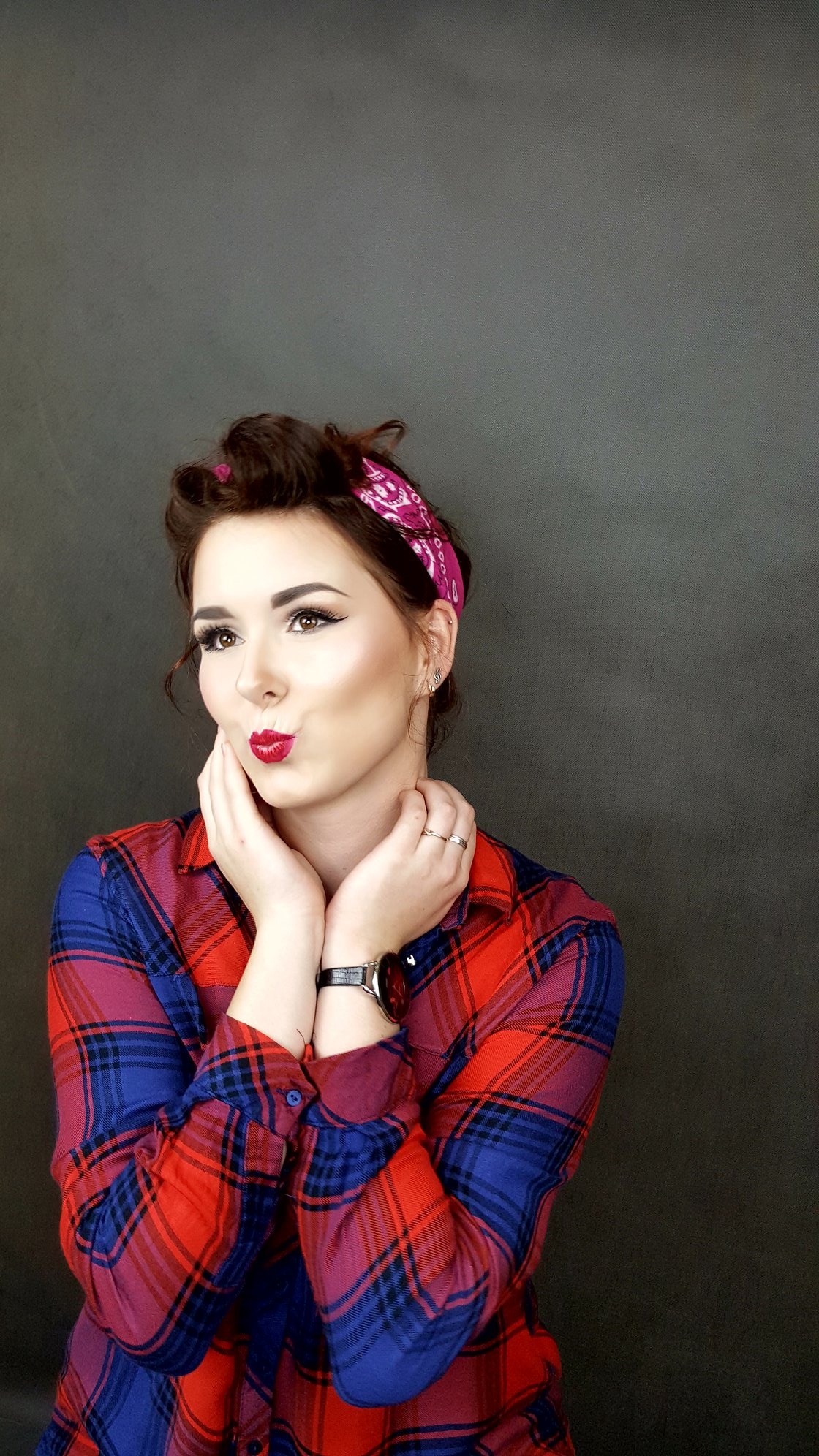 Portret kobiety w stylu pin-up z wyraźnie podkreślonymi ustami i oczami, w koszuli w kratę i z chustą na włosach, na jednolitym szarym tle.