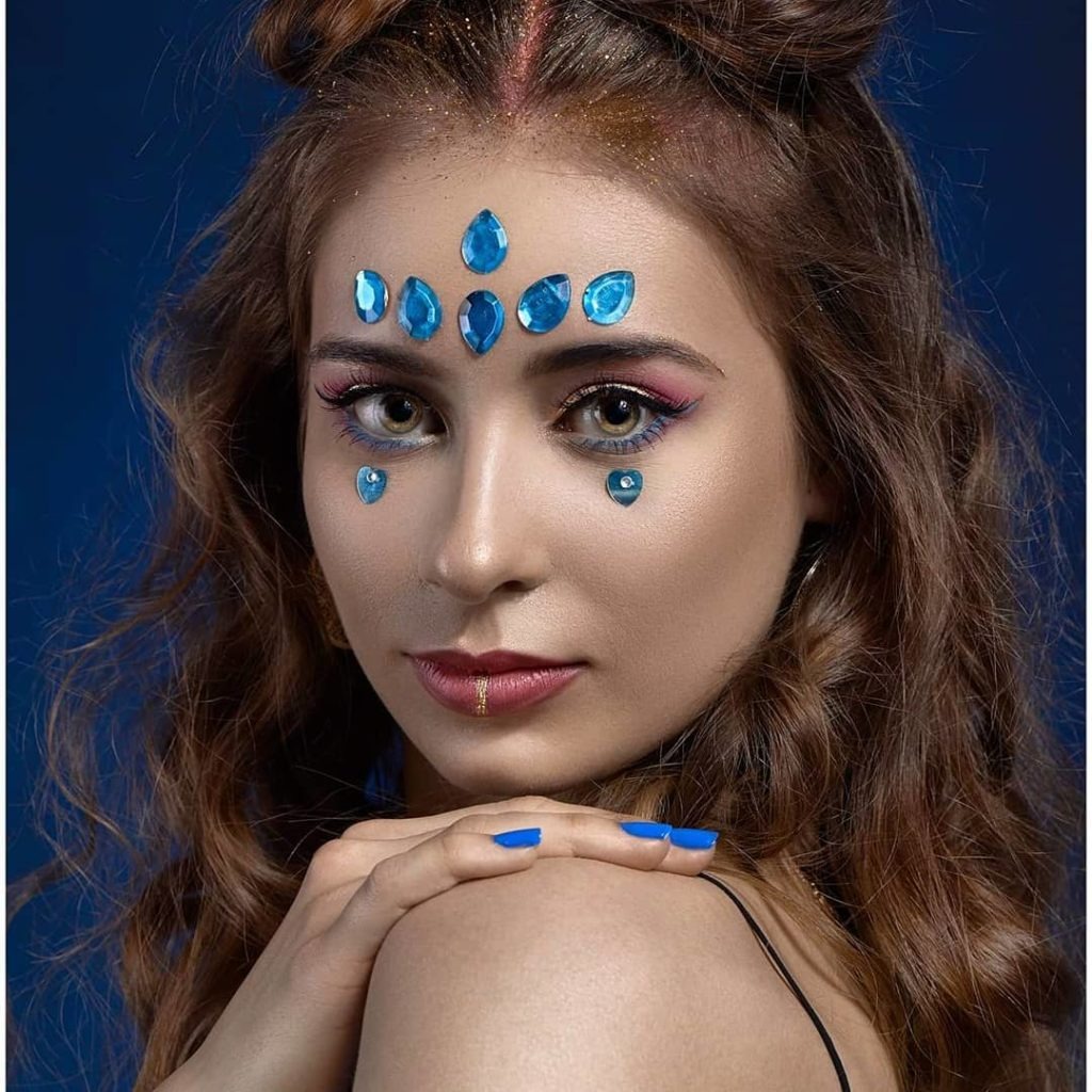 Portret kobiety z artystycznym zdobieniem twarzy: niebieskie kamienie w kształcie łez na czole i pod oczami, różowy cień do powiek, złoty akcent na ustach, niebieski lakier na paznokciach. Włosy...
