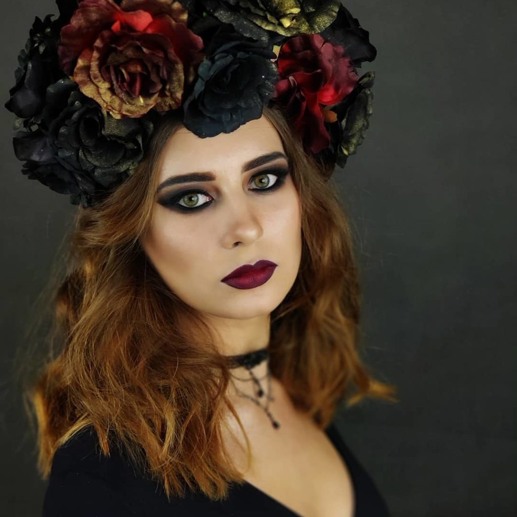 Portret kobiety z intensywnym, gotyckim wizażem: ciemne cienie wokół oczu, bordowe usta i wianek z czarnych i czerwonych róż na głowie, choker na szyi.