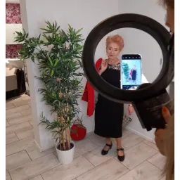 Kobieta w czarnej sukience pozuje do zdjęcia robionego telefonem przez ring light w salonie z rośliną doniczkową i elementami wystroju.