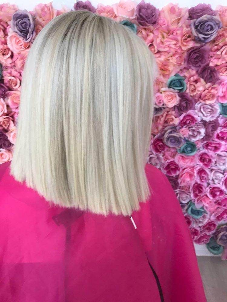 Świeżo ostrzyżone, platynowe blond włosy do ramion, widok z tyłu, na tle ściany z różnokolorowych róż, klientka w różowej pelerynie fryzjerskiej.