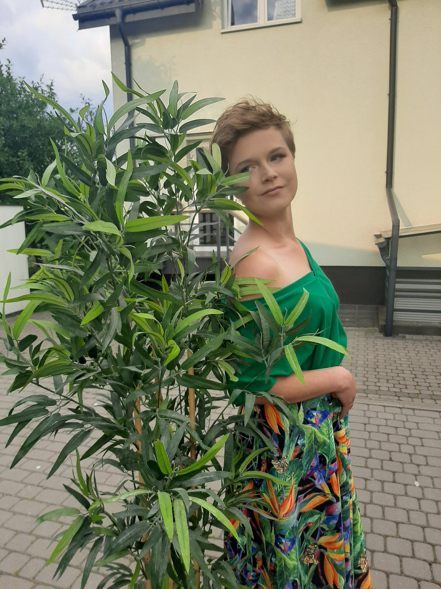Kobieta z krótkimi włosami, w zielonej bluzce i kwiecistej spódnicy, pozuje obok sztucznego drzewka na tle jasnego budynku.