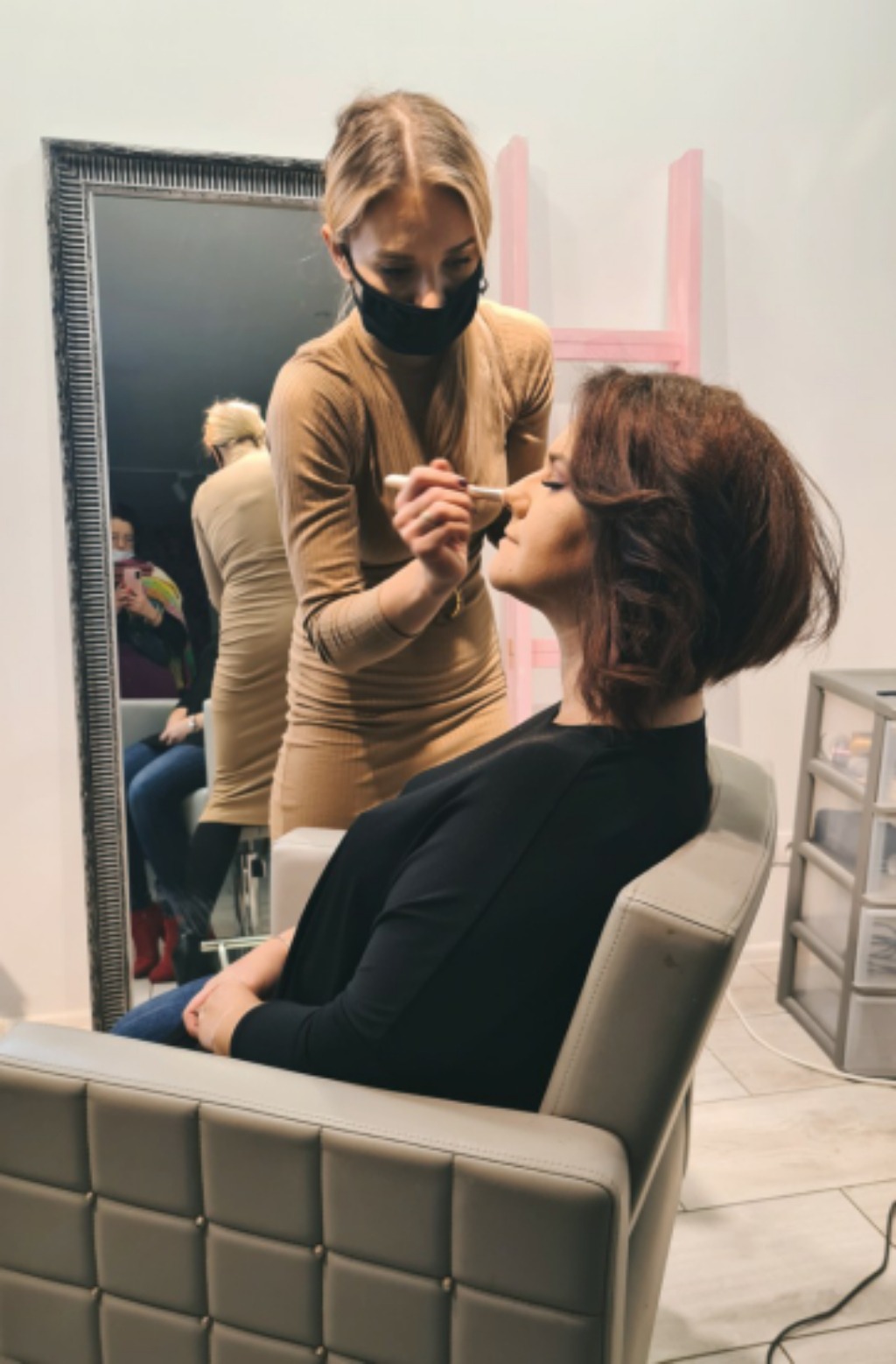 Nakładanie makijażu pędzlem na twarz kobiety siedzącej w fotelu, w tle lustro i różowa drabinka dekoracyjna.