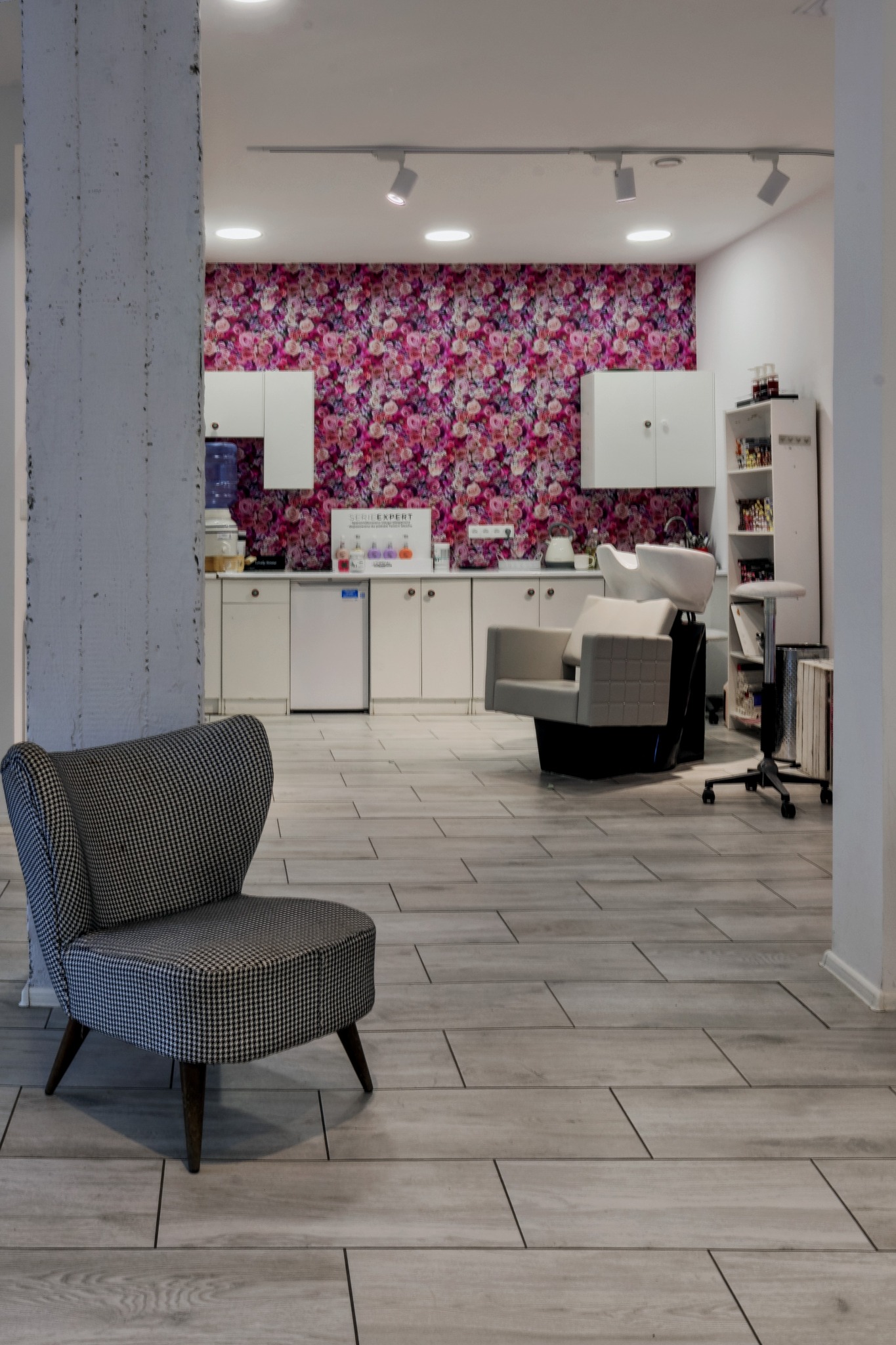 Wnętrze salonu fryzjerskiego z fotelem w pepitkę na pierwszym planie, za nim stanowisko do mycia włosów i ściana w kwiaty.