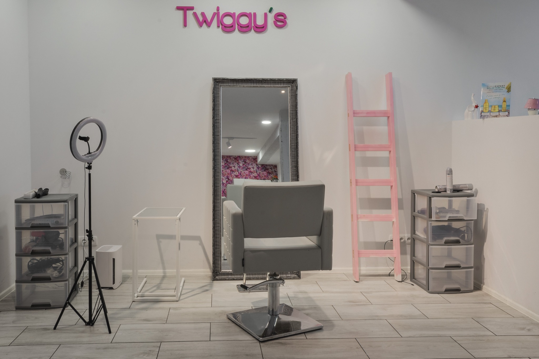 Puste stanowisko w salonie: szare krzesło fryzjerskie, lustro w ozdobnej ramie, ring doświetlający, różowa drabina dekoracyjna, logo 'Twiggy's' na ścianie.