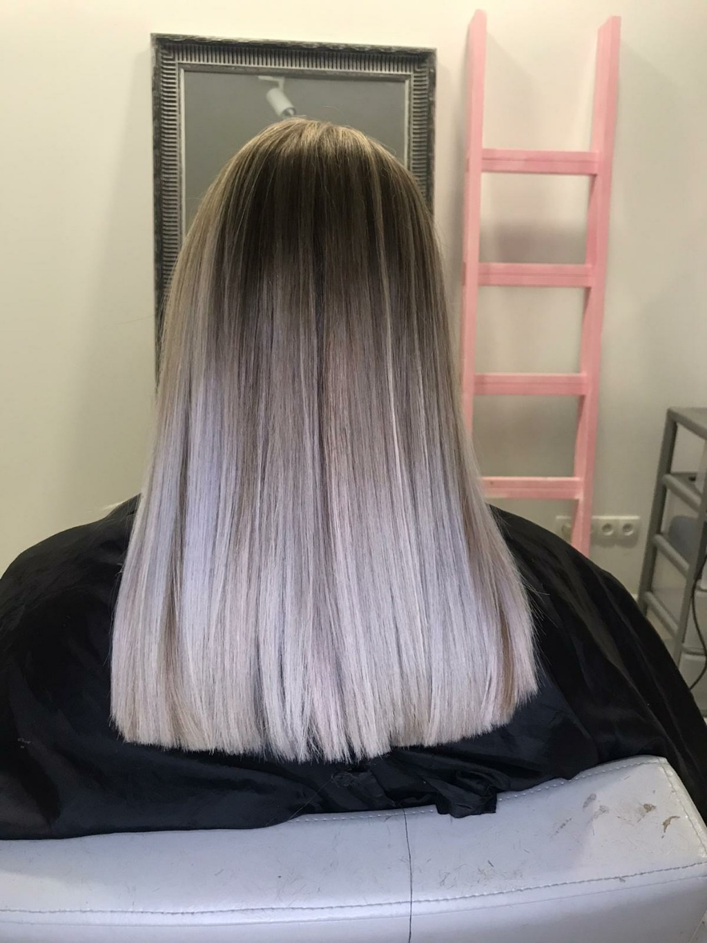 Długie, proste włosy z ombre, od ciemnego brązu u nasady do popielatego blondu na końcach, widoczne z tyłu na tle lustra i różowej drabiny dekoracyjnej w salonie.
